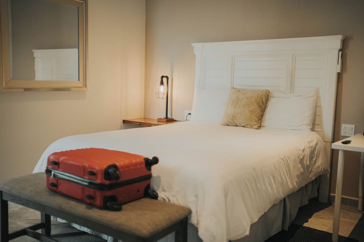 A neatly made bed with a white headboard, a bedside table with a lamp, and a red suitcase on a bench.