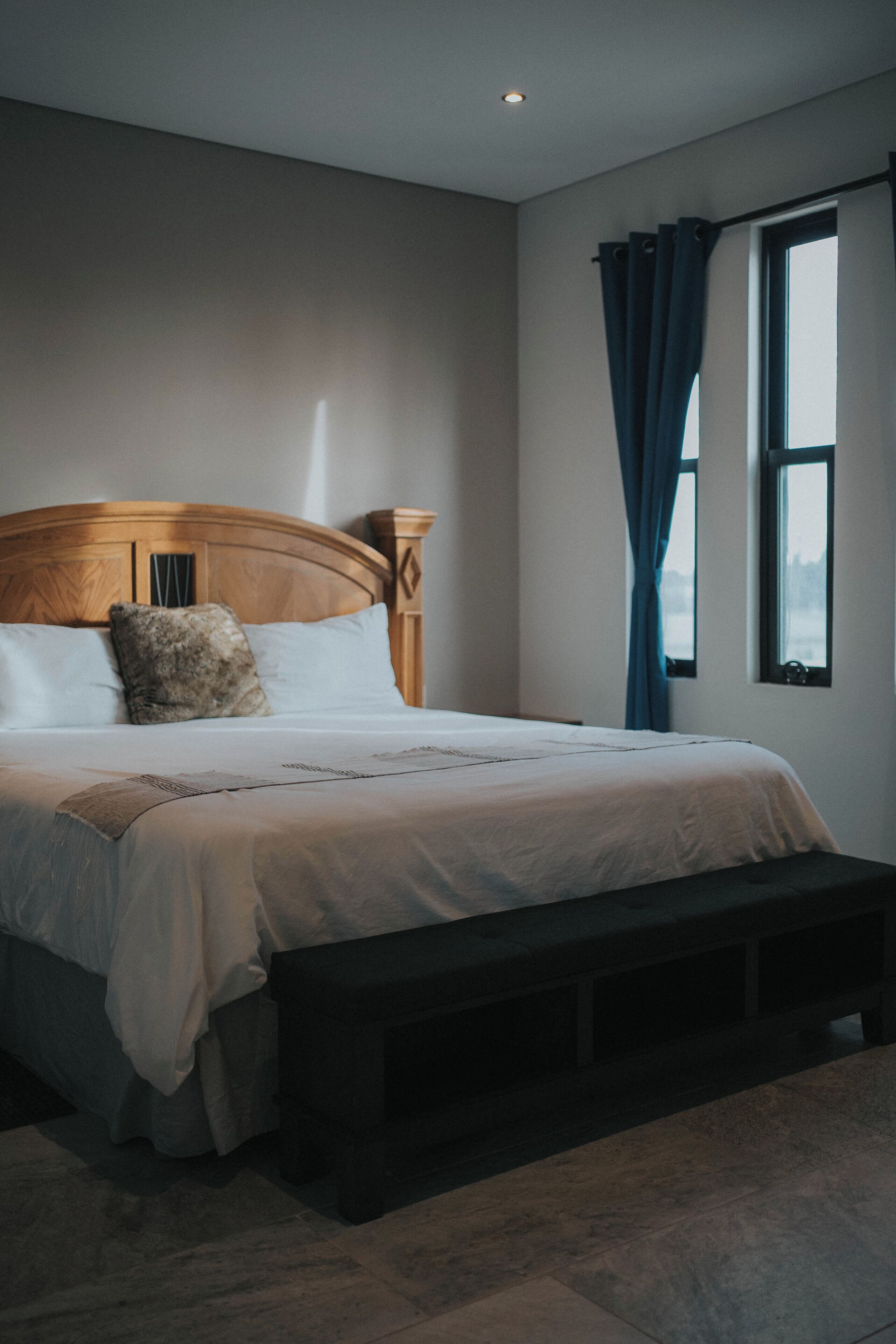 Une chambre à coucher avec une tête de lit en bois, un grand lit recouvert de draps blancs, un banc au pied du lit et une fenêtre ornée de rideaux bleus.