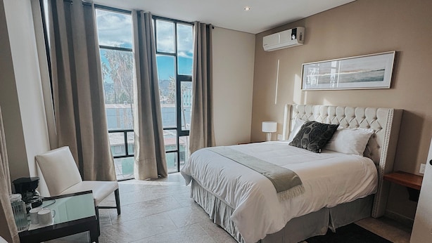 A modern hotel room with a large bed, a desk, and a chair. There is a window with curtains, an air conditioning unit, and a framed picture on the wall. - Suites Lassa (Ensenada)