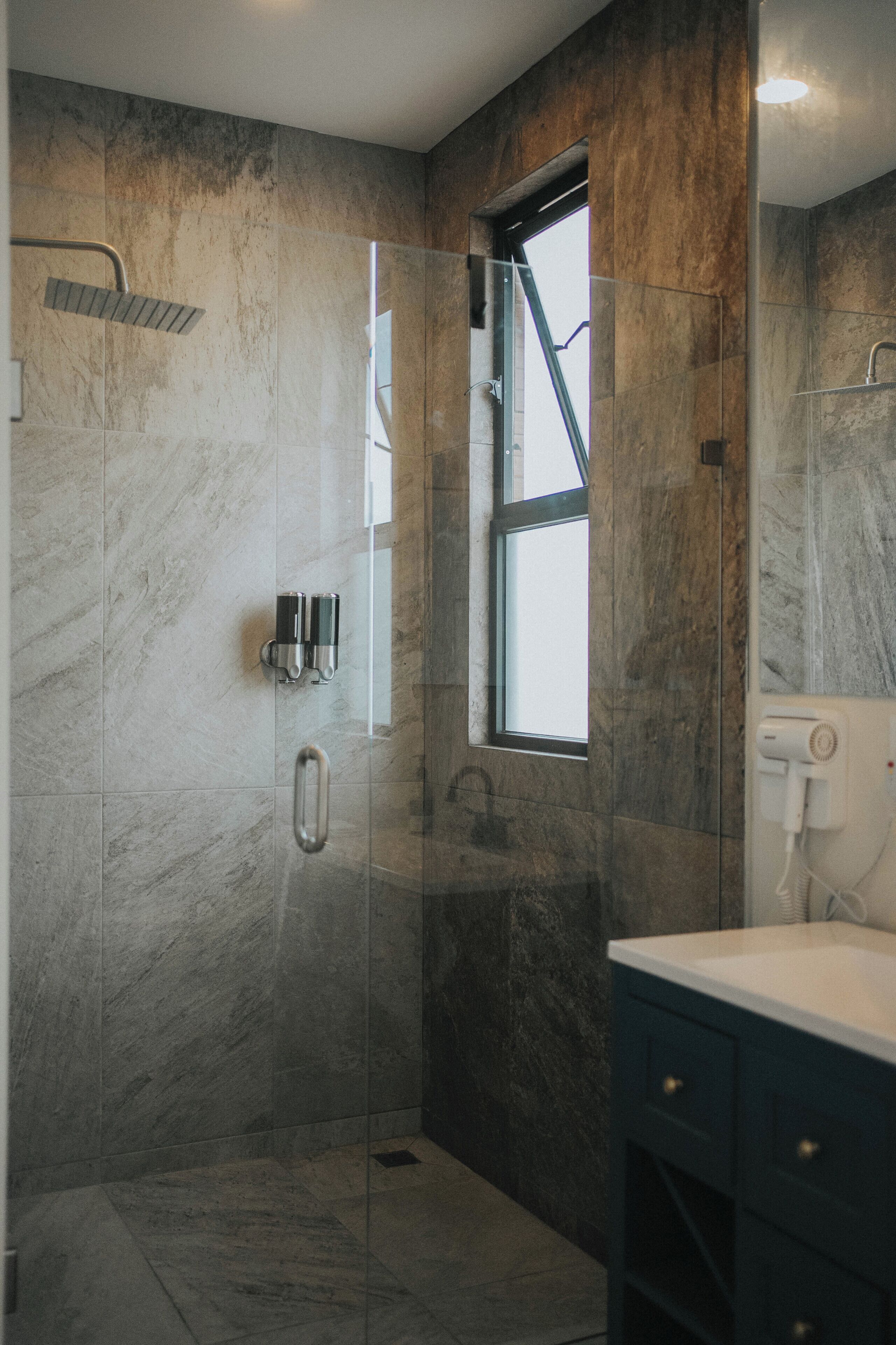 A modern bathroom with a glass-enclosed shower, marble walls, a window, and a built-in sink.