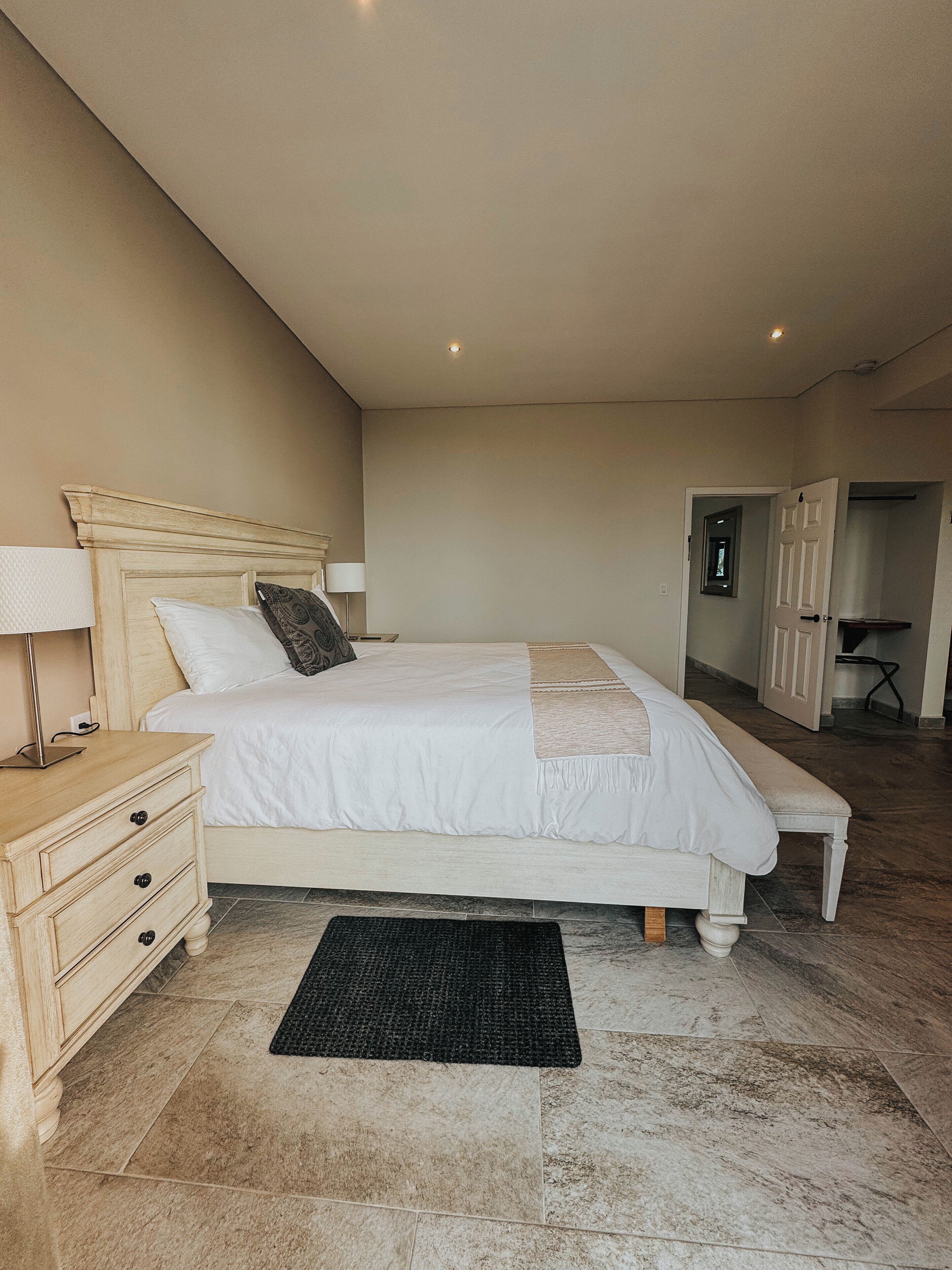 A bedroom with a large bed, a bedside table, a lamp, and a door leading to another room.
