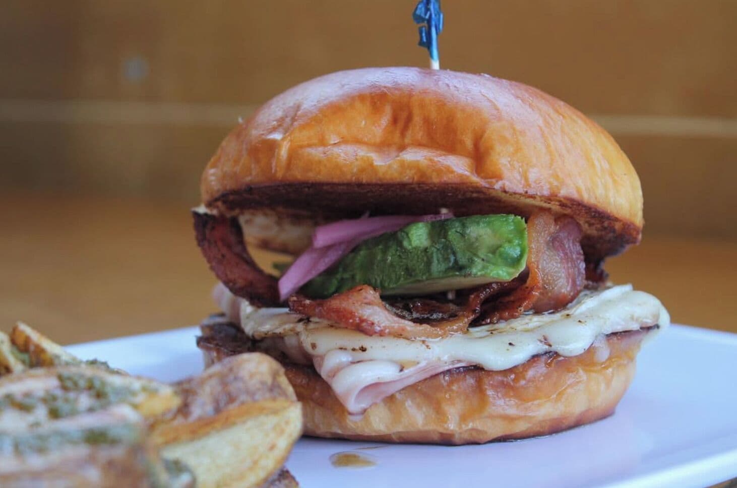 A sandwich with a toothpick, avocado, bacon, and cheese on a bun, accompanied by fries.