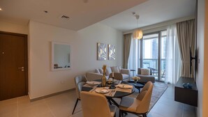 A modern living room with a dining area, a sofa set, a large window with curtains, and a flat-screen TV mounted on the wall. - HomesGetaway-Luxury 1BRApartment in Forte Downtown (Dubai)