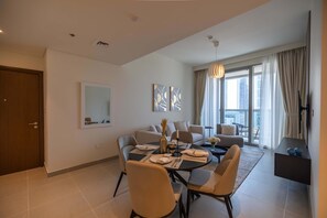 A modern living room with a dining area, a sofa set, a large window with curtains, and a flat-screen TV mounted on the wall.
