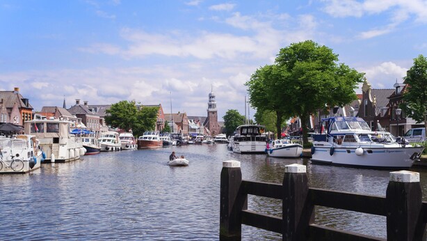 Een gracht met afgemeerde boten, een houten steiger en gebouwen langs het water.