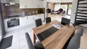 Een moderne keuken met witte kastjes, een inbouwoven en een koffiezetapparaat. Er is een eethoek met een houten tafel en vier stoelen.