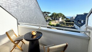 Un balcone con due sedie di legno e un piccolo tavolino rotondo, con vista su una zona residenziale con case e un cielo azzurro intenso.