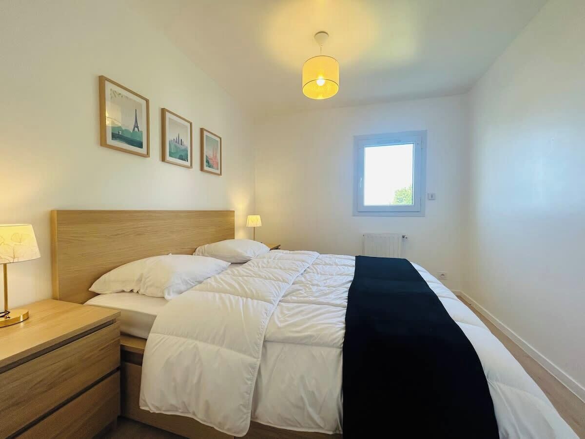 Un lit bien fait, avec une literie blanche et un plaid noir, une table de chevet en bois avec une lampe, et deux tableaux encadrés accrochés au mur.