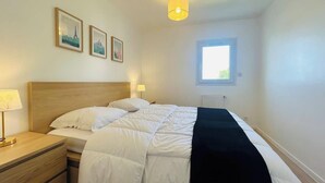 En pænt redt seng med hvide sengetæpper og et sort pled, et trænatbord med en lampe, samt to billeder i rammer på væggen.