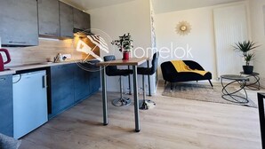 Et moderne køkken med lysegrå skabe, et lille køleskab og et spiseområde med bord og stole. Der er en sort sofa og et lille rundt bord med en plante.