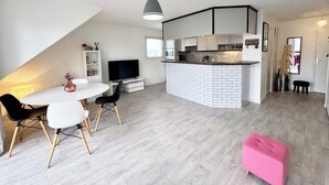 Un espacio moderno con comedor, cocina y un pequeño rincón de descanso con un pouf rosa.
