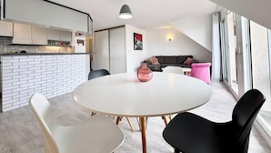 Una cocina moderna con armarios blancos y encimera blanca. Hay una zona de comedor con una mesa redonda y sillas. Un jarrón rosa está sobre la mesa.