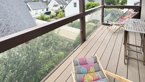 Terraza de madera con una silla de lounge y una mesa, con vistas a una zona residencial con casas y vegetación.