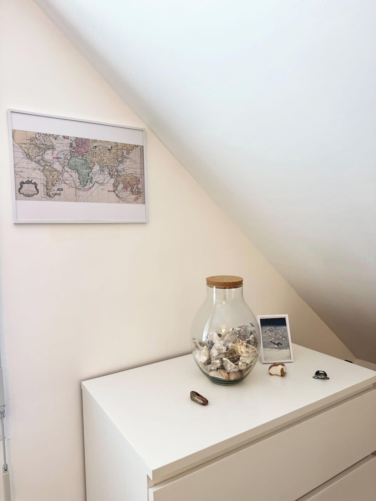 A white desk with a glass jar containing seashells, a framed map, and a small framed picture.