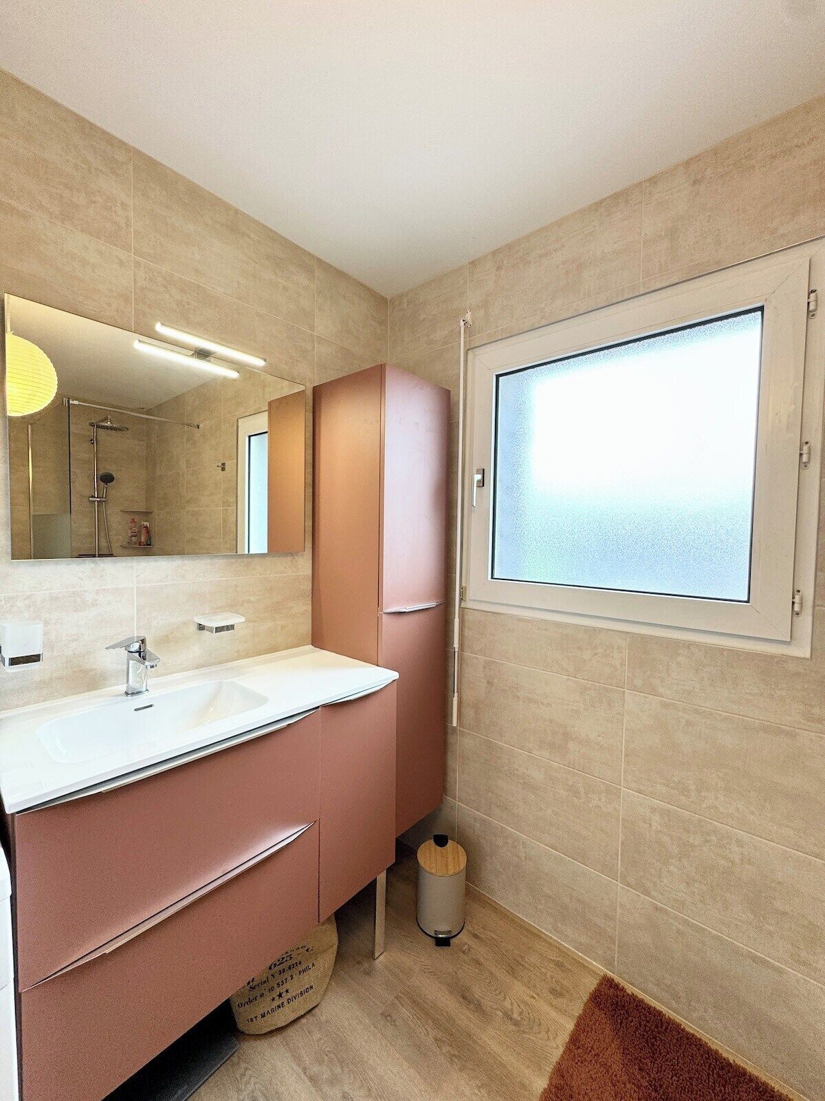 Uma casa de banho moderna com um espelho grande, uma sanita branca, um armário cor-de-rosa e uma janela com vidro jateado.