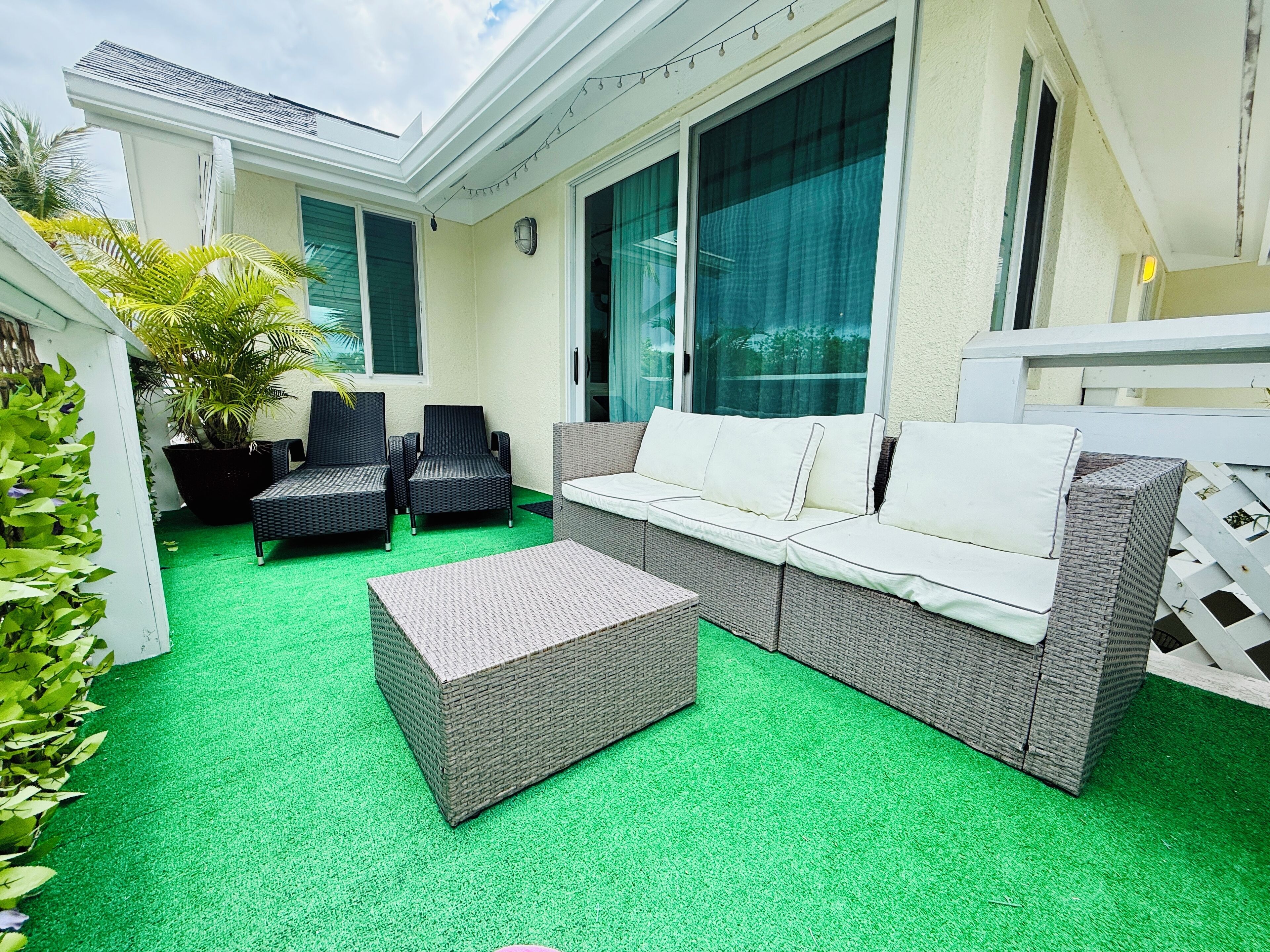 A patio with a grey sofa, two black chairs, and a grey ottoman. There are potted plants and a string of lights.