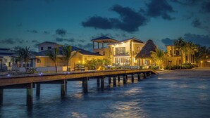 A beachfront property with a wooden pier extending into the water, a pool area with lounge chairs, and a thatched-roof structure.