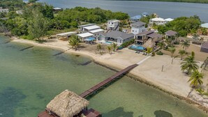 An aerial view of a coastal property with a beach, a thatched-roof structure on stilts, and several houses with a pool.