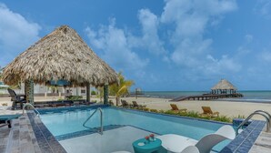 A pool area with a thatched-roof structure, lounge chairs, and a view of the beach and ocean.