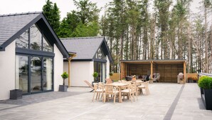 A modern house with a patio area featuring a wooden table and chairs, surrounded by tall trees. - Dreamy 5-Bedroom Cottage with Private Hot Tub (Aberyswyth)