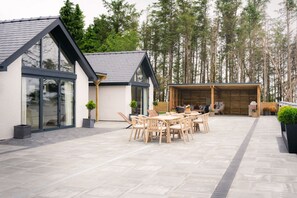 Et moderne hus med en patio med et træbord og stole, omgivet af høje træer.