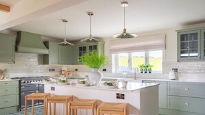 A kitchen with green cabinets, a white island with stools, a stove, and a window with a view of the outdoors. - Dreamy 5-Bedroom Cottage with Private Hot Tub (Aberyswyth)