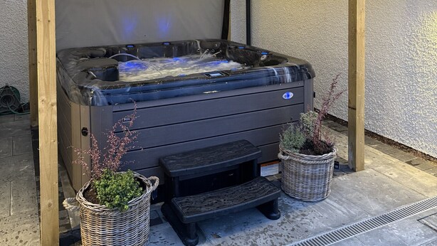 A hot tub under a wooden pergola with string lights, surrounded by potted plants and a stone patio.