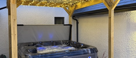 A hot tub under a wooden pergola with string lights, surrounded by potted plants and a stone patio.