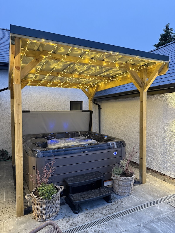 A hot tub under a wooden pergola with string lights, surrounded by potted plants and a stone patio.