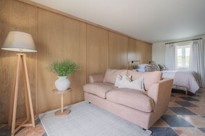 A cozy living room with a beige sofa, a small side table with a plant, a floor lamp, and a view of a bedroom with a bed and curtains.