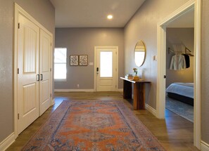 A hallway with a patterned rug, a door, a console table with a mirror, and a bed in the adjacent room. - Atlas Cottage Cozy Stay for 2 (Dripping Springs)