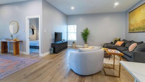 Un salon moderne avec un canapé gris, un pouf rond, un téléviseur à écran plat et une grande fenêtre laissant entrer la lumière naturelle.