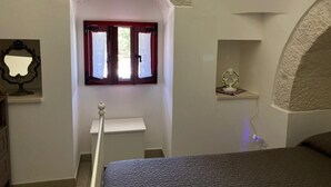 A bedroom with a bed, a window with red shutters, a small table with a decorative item, and a wall-mounted shelf.