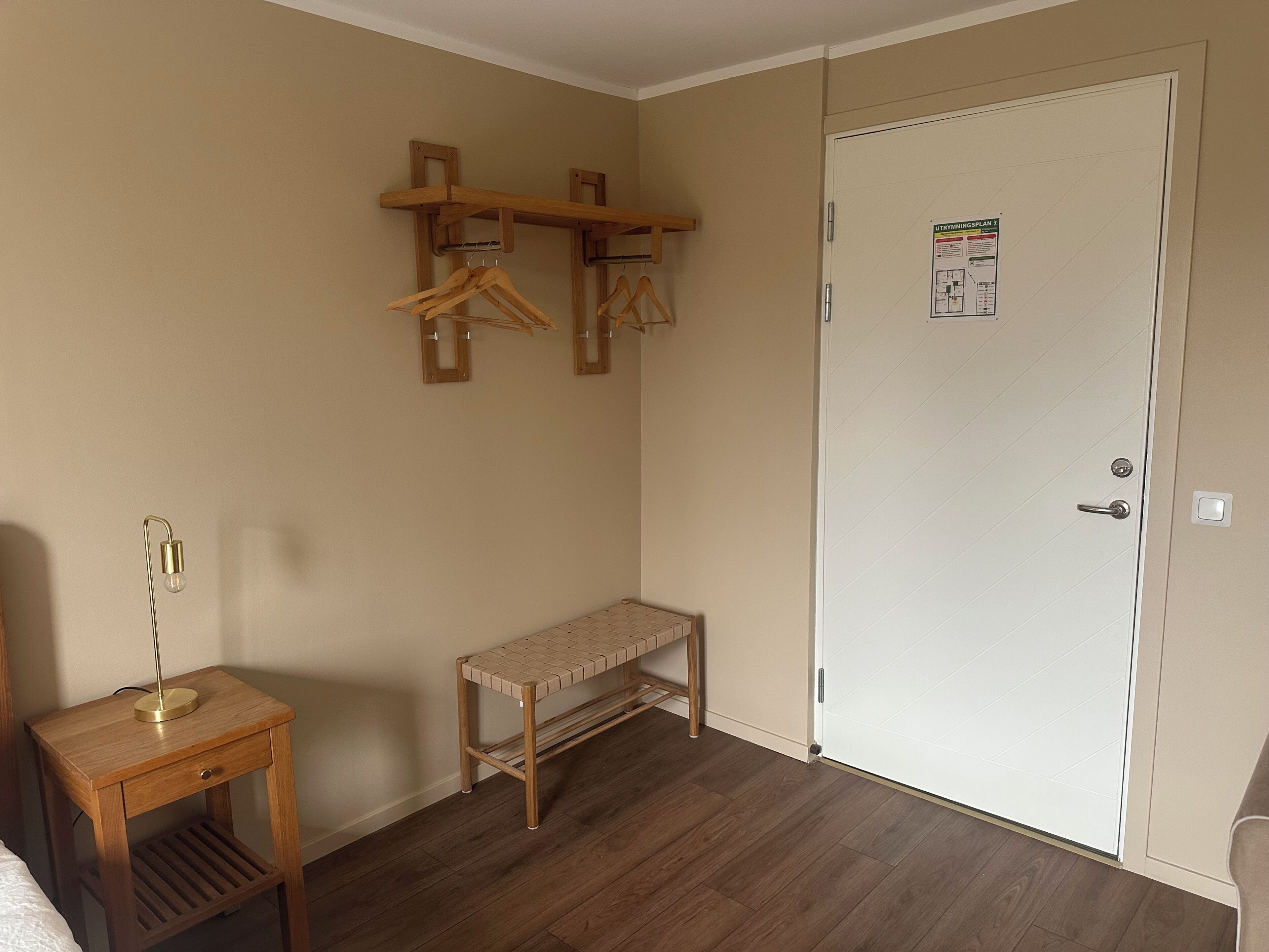 Una habitación con un banco de madera, una mesita con una lámpara, un perchero y una puerta con un aviso.