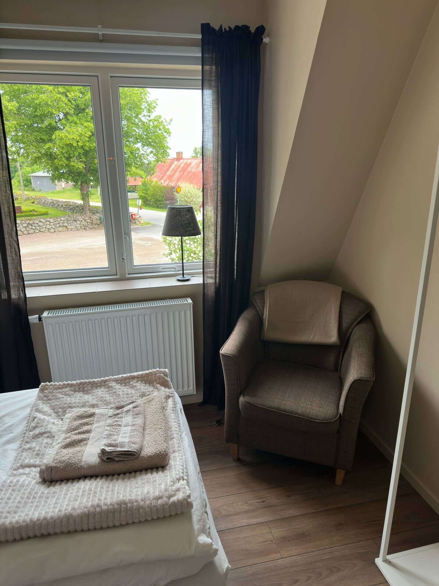 A bedroom with a bed, a chair, a window with a view of greenery and a building, and a radiator.
