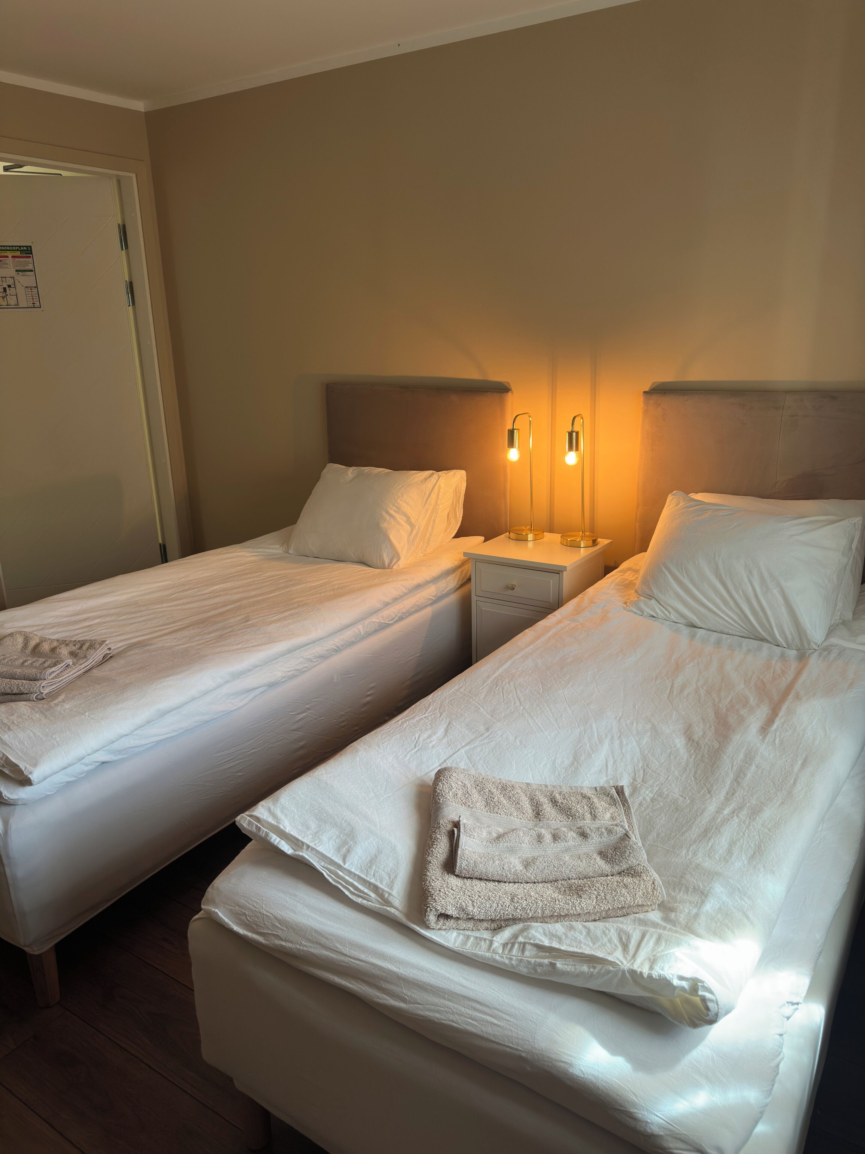 A hotel room with two single beds, a bedside table with a lamp, a door, and a wall-mounted light fixture.