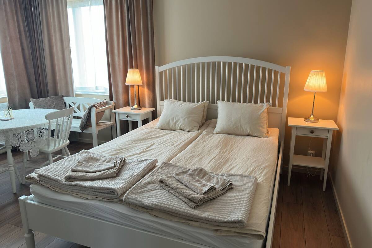 A neatly made bed with pillows, two bedside tables with lamps, a small table with a chair, and a window with curtains.