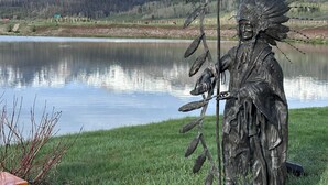 Eine Statue einer indianischen Figur, die einen Bogen und Pfeil hält, an einem See mit Bergen im Hintergrund.
