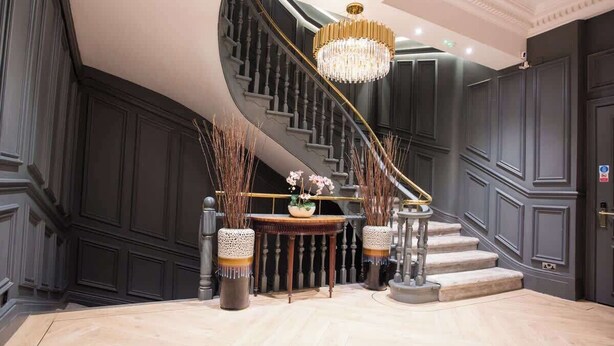 A grand staircase with a chandelier, dark wood paneling, and a round table with decorative plants.