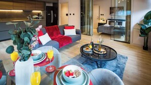 A modern living room with a sofa, coffee table, and dining area. There is a kitchen in the background with a refrigerator and cabinets. - Stylish City Escape | Walk to Chinatown | AC inc. (Greater Manchester)