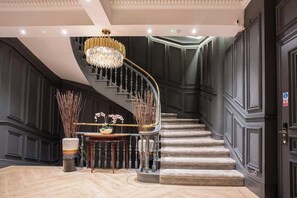 Un magnifique escalier orné d’un lustre, des boiseries sombres et une petite table décorée de fleurs.
