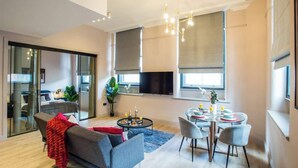 A modern living room with a grey sofa, a dining table with chairs, a TV mounted on the wall, and large windows with blinds. - Stylish City Escape | Walk to Chinatown | AC inc. (Greater Manchester)