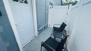 Un pequeño espacio al aire libre cercado, con una puerta y una valla blancos. Hay dos sillas negras y una mesa pequeña.