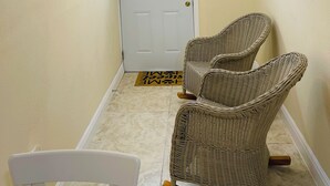 Un pasillo con una puerta blanca, dos sillas de mimbre, una lámpara en el techo y un cuadro en la pared.