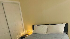 Una cama bien hecha con una colcha azul y almohadas blancas, una toalla blanca doblada sobre la cama y una lámpara de noche en una mesita.