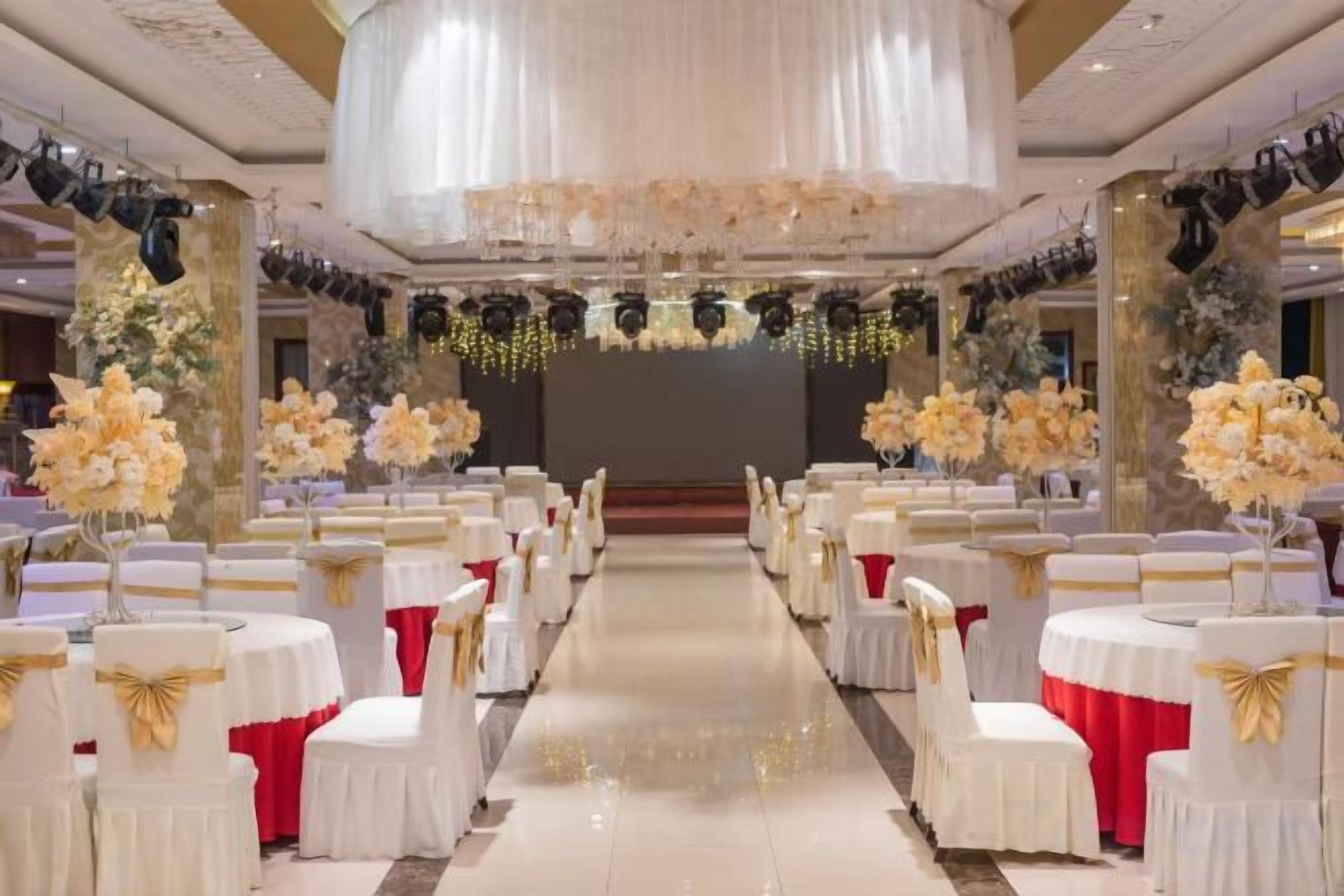 A banquet hall with rows of tables and chairs, a stage at the front, and floral decorations.