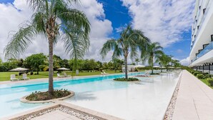 A pool area with multiple swimming pools, palm trees, lounge chairs, and a modern building in the background.
