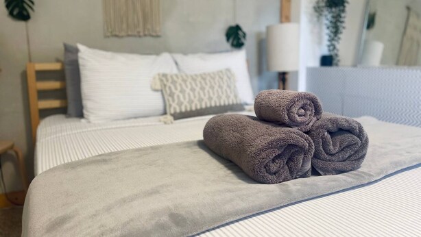 Una cama bien tendida con toallas enrolladas, un cabecero de madera, un cojín estampado y una planta en la pared.