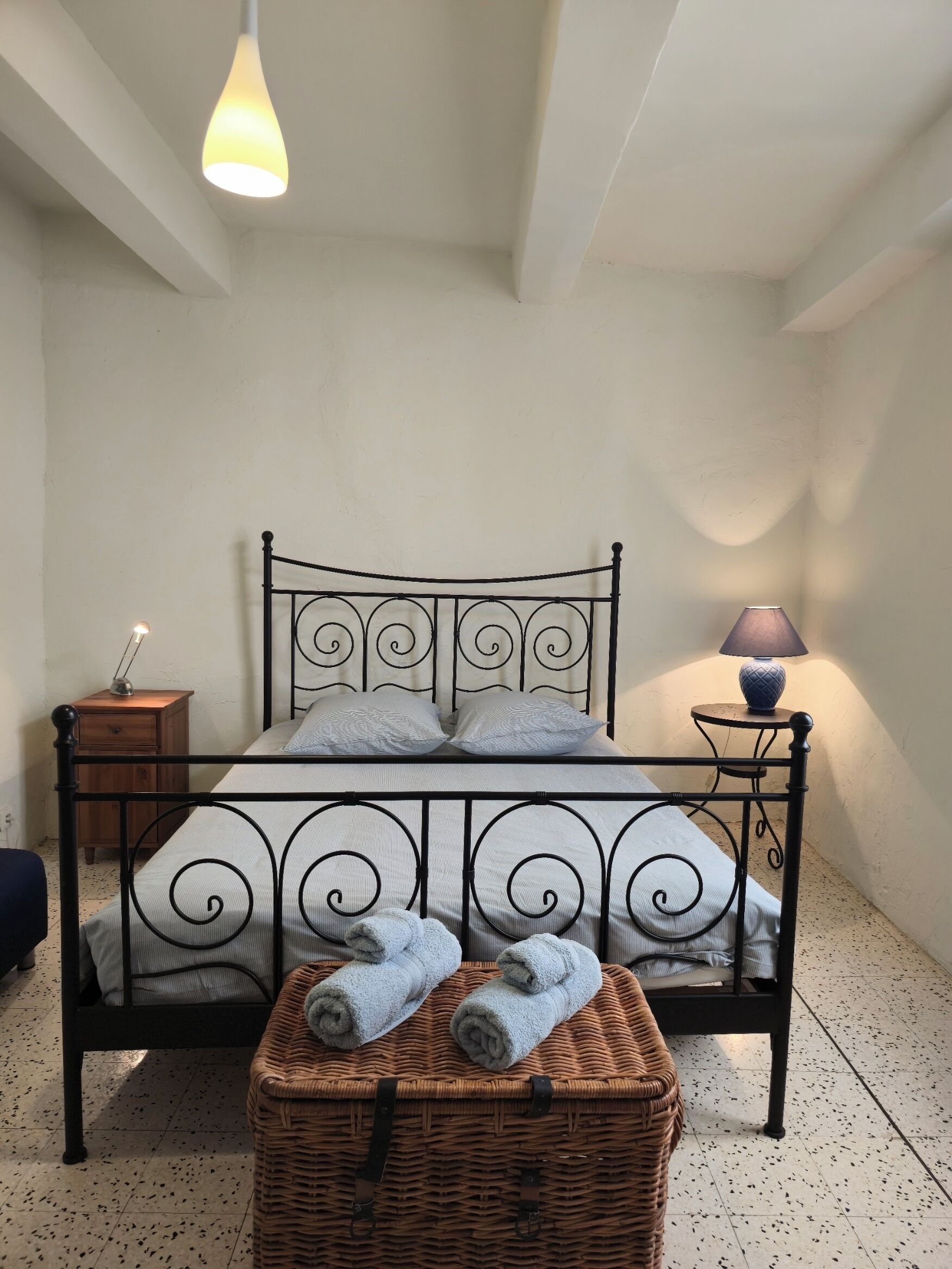 A neatly arranged bedroom with a metal-framed bed, a bedside table, a lamp, and a wicker basket with towels.