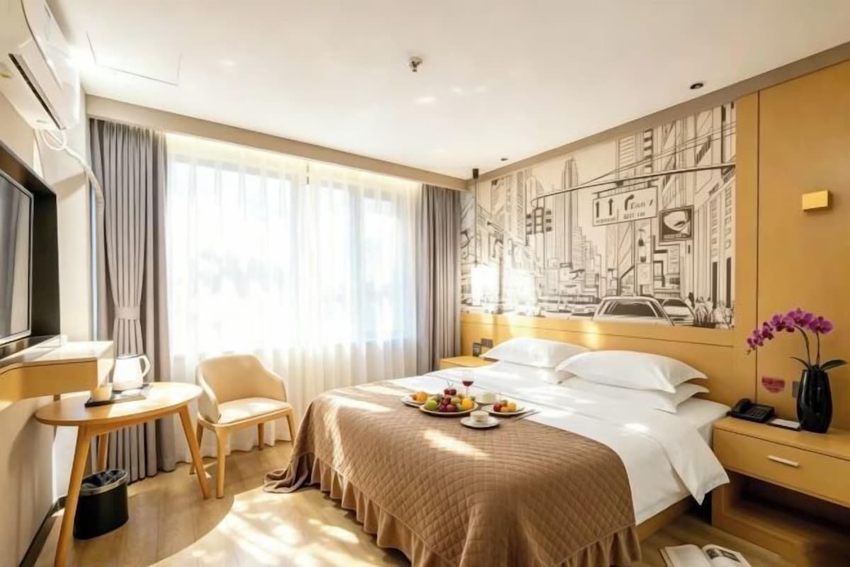 Habitación de hotel con cama, escritorio, silla, televisor y ventana con cortinas, además de un mural con vista a la ciudad.